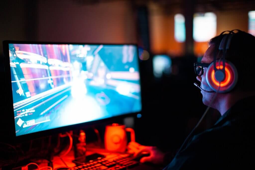 A gamer wearing glowing orange headphones plays a fast-paced 3D game on a widescreen monitor in a dark room.