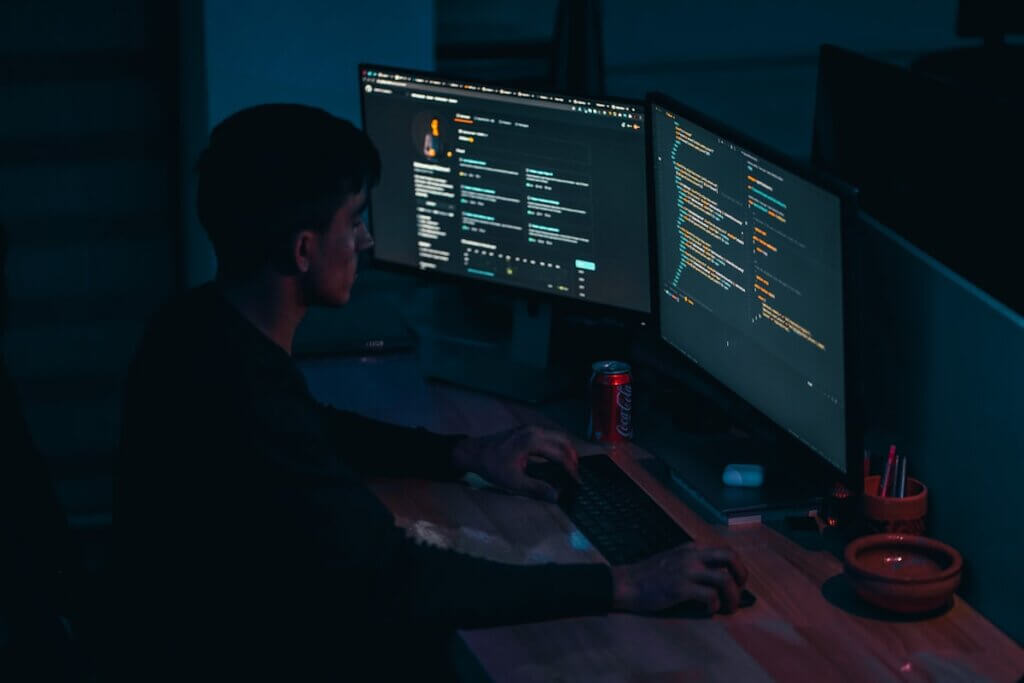 A solo developer codes at a dark dual-monitor workstation with colorful AI-assisted code on screen.