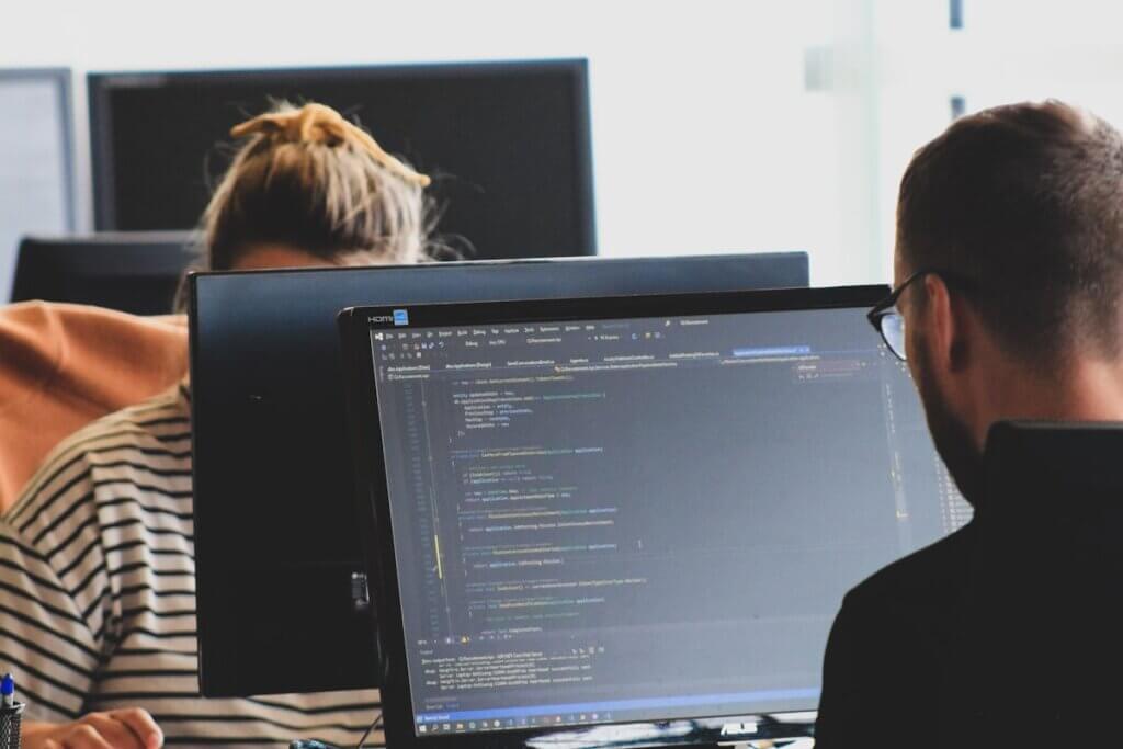 Two developers review code across from each other on ASUS monitors in a bright office, collaborating on a software project.
