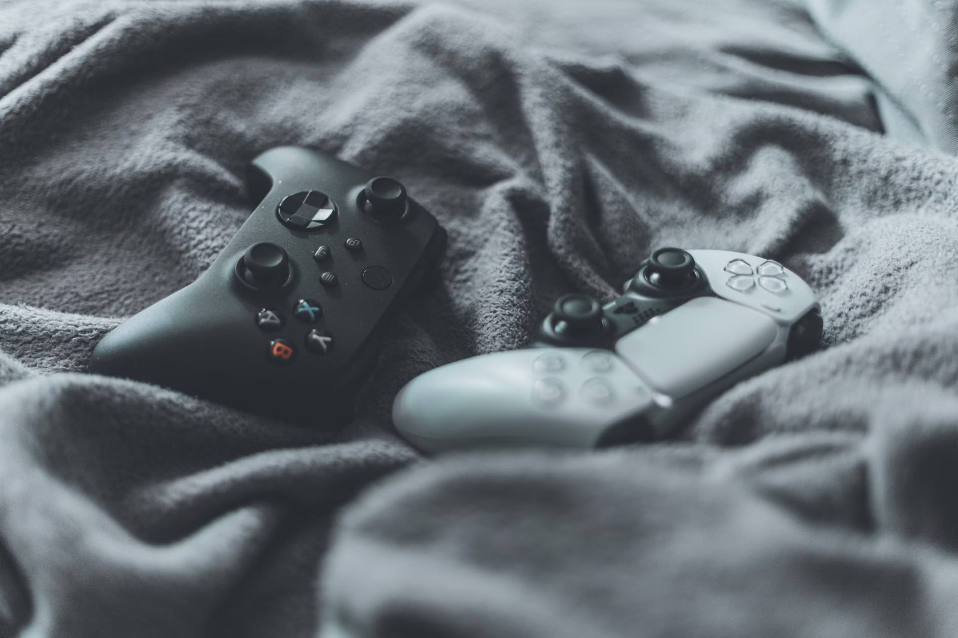 Black Xbox One game controller and Playstation Dualsense Edge controller resting on a dark surface, viewed from above.