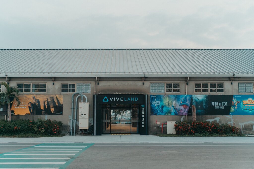 Exterior of the VIVELAND warehouse-style building with a glass entrance and VR-themed banners, including artwork for "Alice" and "House of Fear."